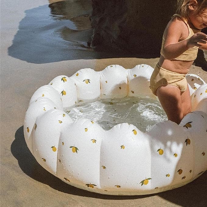 Child playing in a small inflatable pool with lemon pattern on a sandy beach.